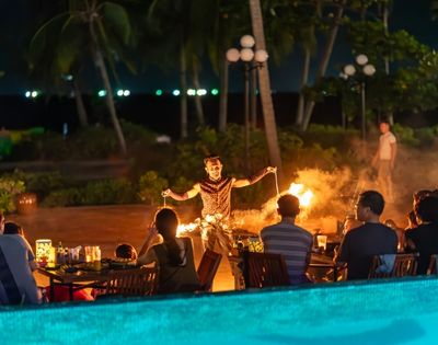 A fire performer entertains a seated audience outdoors at night. Palm trees and lanterns enhance the tropical ambiance. Warm lighting creates an exciting mood.