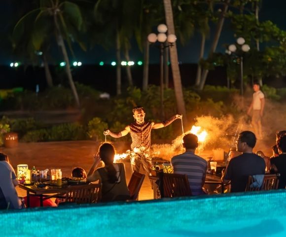 A fire performer entertains a seated audience outdoors at night. Palm trees and lanterns enhance the tropical ambiance. Warm lighting creates an exciting mood.