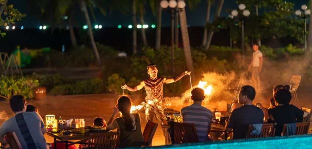 A fire performer entertains a seated audience outdoors at night. Palm trees and lanterns enhance the tropical ambiance. Warm lighting creates an exciting mood.
