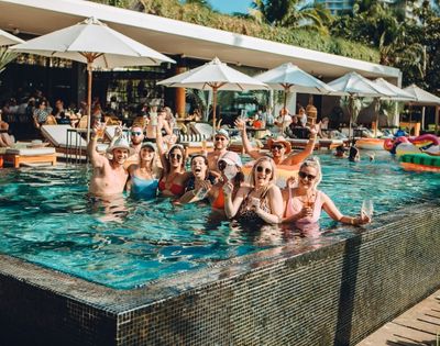A group of friends in swimwear stands in a vibrant pool, smiling and raising drinks. Umbrellas and palm trees suggest a tropical, relaxed atmosphere.