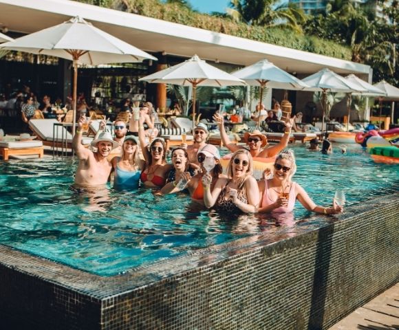A group of friends in swimwear stands in a vibrant pool, smiling and raising drinks. Umbrellas and palm trees suggest a tropical, relaxed atmosphere.