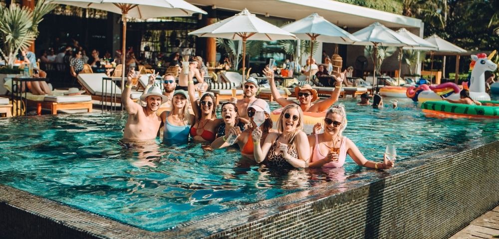 A group of friends in swimwear stands in a vibrant pool, smiling and raising drinks. Umbrellas and palm trees suggest a tropical, relaxed atmosphere.