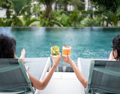 Two people relax on poolside loungers, holding colorful cocktails. The background features lush green foliage, suggesting a peaceful, tropical setting.