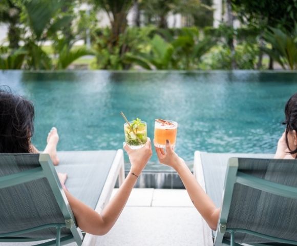 Two people relax on poolside loungers, holding colorful cocktails. The background features lush green foliage, suggesting a peaceful, tropical setting.