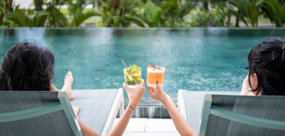 Two people relax on poolside loungers, holding colorful cocktails. The background features lush green foliage, suggesting a peaceful, tropical setting.
