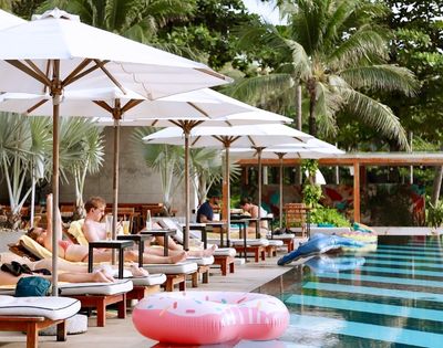 People relax on poolside loungers under white umbrellas, surrounded by palm trees. Inflatable floats are in the pool, creating a tropical, serene atmosphere.