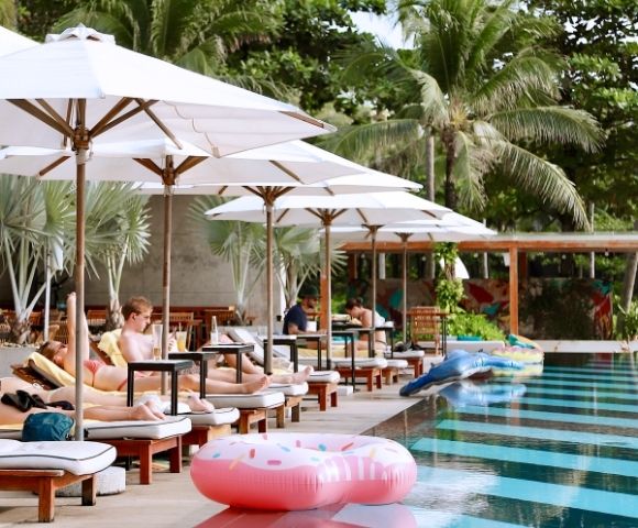 People relax on poolside loungers under white umbrellas, surrounded by palm trees. Inflatable floats are in the pool, creating a tropical, serene atmosphere.
