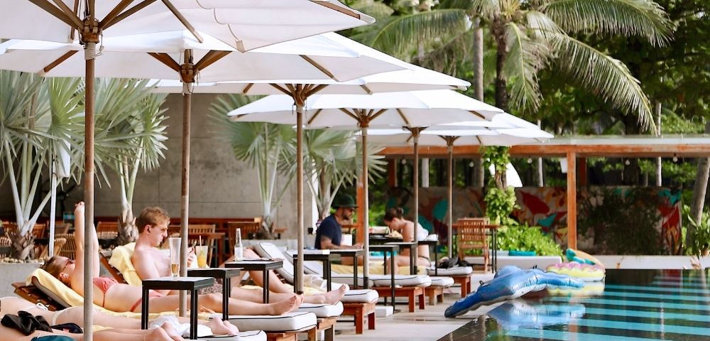 People relax on poolside loungers under white umbrellas, surrounded by palm trees. Inflatable floats are in the pool, creating a tropical, serene atmosphere.
