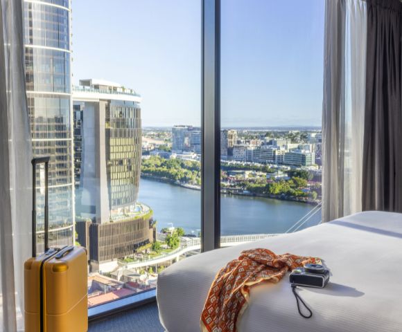 Bright hotel room with city view. Large window reveals river and skyscrapers. Bed with camera and scarf. Yellow suitcase next to window.