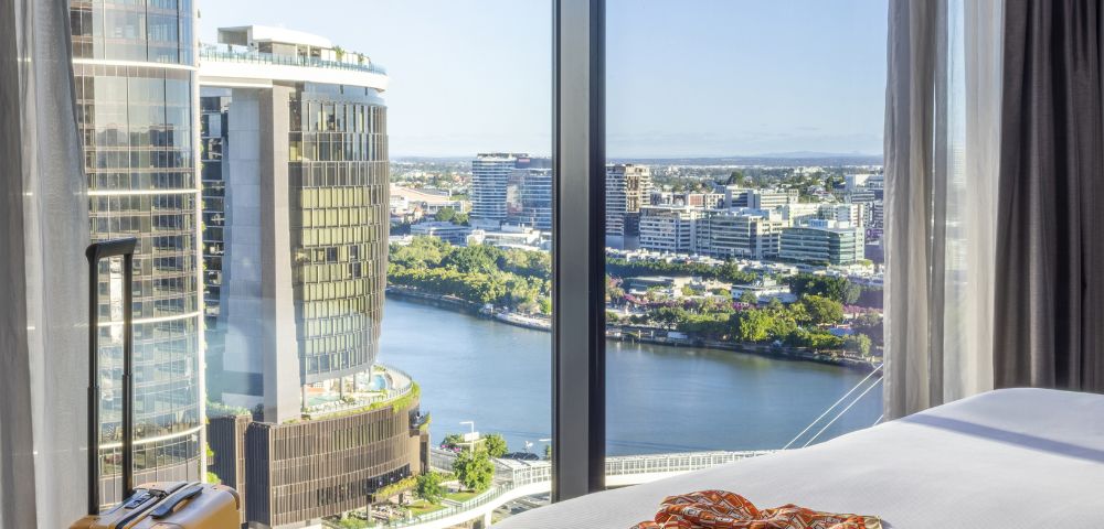 Bright hotel room with city view. Large window reveals river and skyscrapers. Bed with camera and scarf. Yellow suitcase next to window.