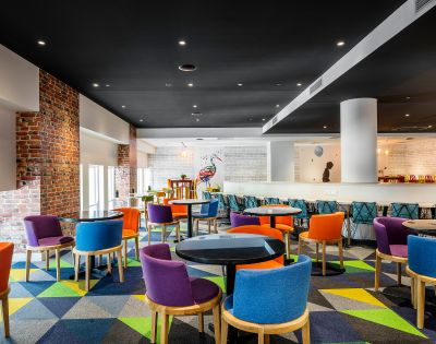 Vibrant, modern lounge with colorful chairs around black tables on a geometric-patterned carpet. Exposed brick and a mural add an artistic touch.
