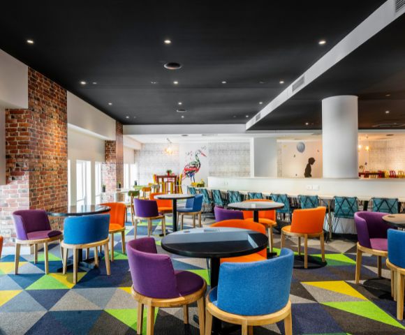 Vibrant, modern lounge with colorful chairs around black tables on a geometric-patterned carpet. Exposed brick and a mural add an artistic touch.