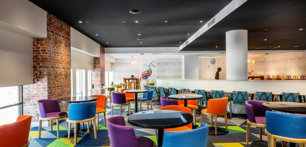 Vibrant, modern lounge with colorful chairs around black tables on a geometric-patterned carpet. Exposed brick and a mural add an artistic touch.