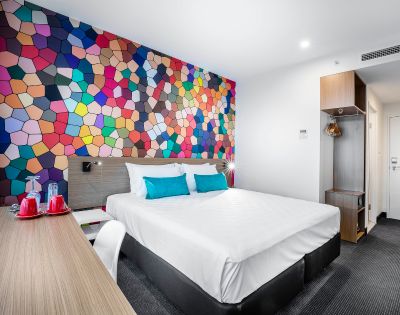 A modern hotel room features a vibrant, geometric mosaic accent wall. The room has a white bed with blue pillows, a wooden desk, and warm lighting, creating a lively yet cozy atmosphere.