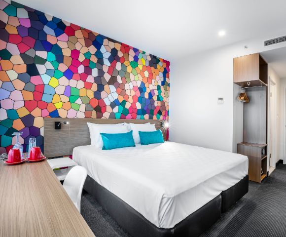 A modern hotel room features a vibrant, geometric mosaic accent wall. The room has a white bed with blue pillows, a wooden desk, and warm lighting, creating a lively yet cozy atmosphere.