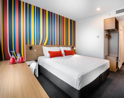 Modern hotel room with a vibrant multicolored striped wall. Features a neatly made double bed with red and white pillows. A sleek desk with red mugs and water glasses adds a cozy, inviting feel.