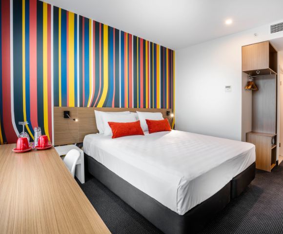 Modern hotel room with a vibrant multicolored striped wall. Features a neatly made double bed with red and white pillows. A sleek desk with red mugs and water glasses adds a cozy, inviting feel.