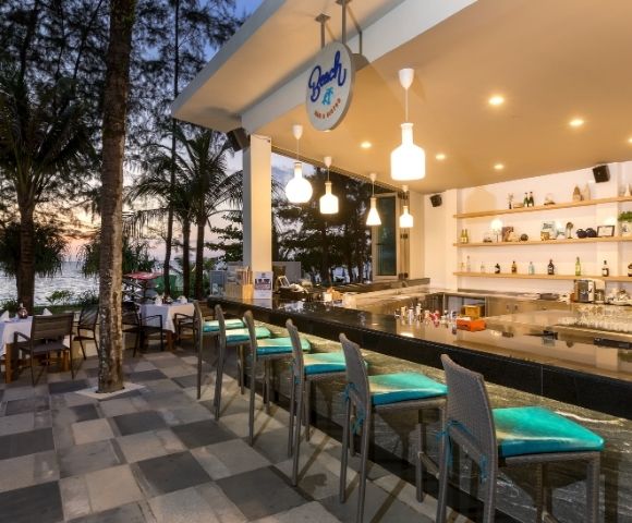 Outdoor bar with turquoise-cushioned stools, overlooking a sunset beach. Warm lighting, bottles on shelves, and trees create a relaxed, inviting atmosphere.