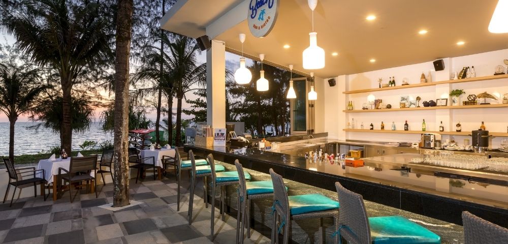Outdoor bar with turquoise-cushioned stools, overlooking a sunset beach. Warm lighting, bottles on shelves, and trees create a relaxed, inviting atmosphere.