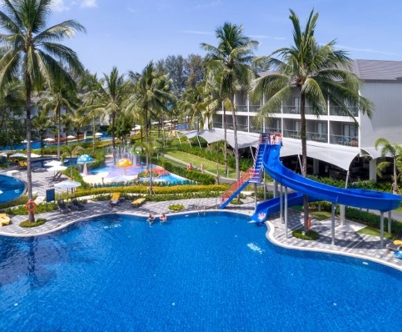 Resort pool area with lush palm trees, a vibrant blue slide, and sunlit lounge chairs. A relaxing, tropical atmosphere under a clear blue sky.