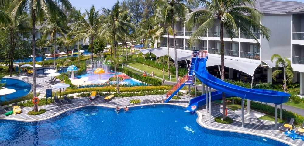Resort pool area with lush palm trees, a vibrant blue slide, and sunlit lounge chairs. A relaxing, tropical atmosphere under a clear blue sky.
