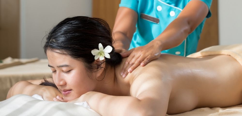A woman with a flower in her hair receives a relaxing back massage from a masseuse wearing a teal uniform. The setting conveys calm and serenity.