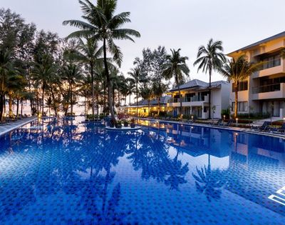 Luxurious resort pool at dusk, surrounded by palm trees and modern buildings. The calm blue water reflects soft evening lights, creating a serene ambiance.