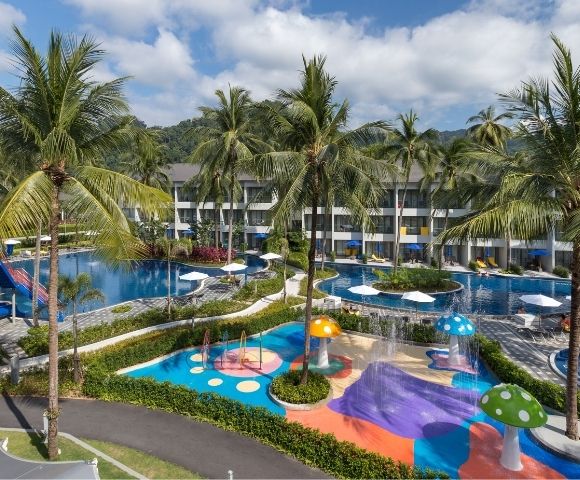 Tropical resort with two pools surrounded by palm trees. Colorful water play area for children with slides and mushroom fountains. Relaxing atmosphere.