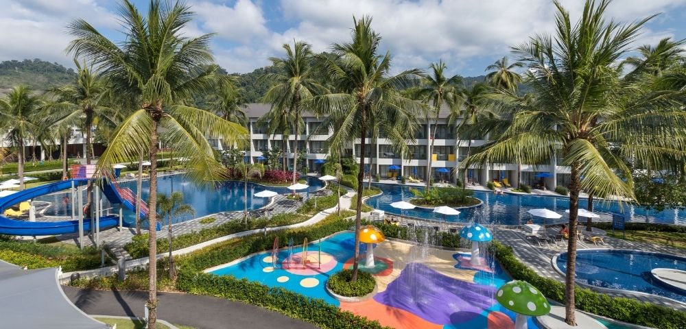 Tropical resort with two pools surrounded by palm trees. Colorful water play area for children with slides and mushroom fountains. Relaxing atmosphere.