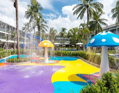Colorful splash pad with mushroom fountains, surrounded by palm trees and modern buildings. Vibrant and playful atmosphere under a bright blue sky.