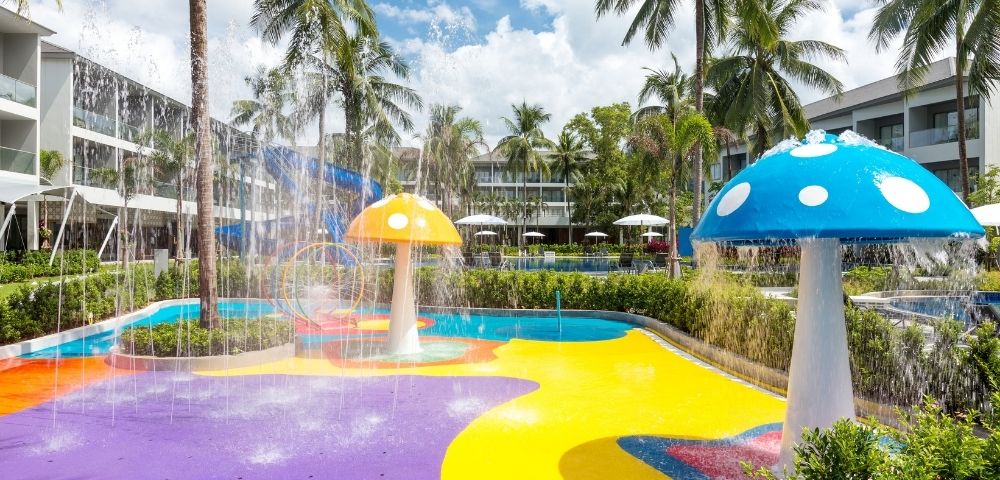 Colorful splash pad with mushroom fountains, surrounded by palm trees and modern buildings. Vibrant and playful atmosphere under a bright blue sky.