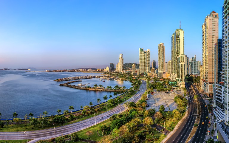 A panoramic view of a coastal city at sunset, showcasing tall skyscrapers, a curving shoreline, and palm-lined roads. The scene conveys a serene urban atmosphere.