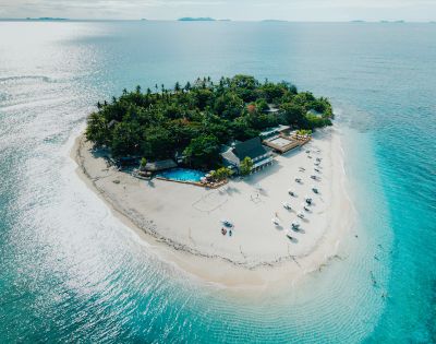 Aerial view of a small tropical island with lush greenery, surrounded by turquoise water. White sandy beach features sun loungers and a poolside building. Relaxing vibe.