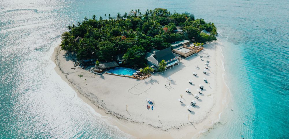 Aerial view of a small tropical island with lush greenery, surrounded by turquoise water. White sandy beach features sun loungers and a poolside building. Relaxing vibe.