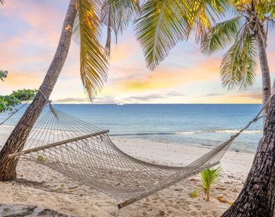 A hammock hangs between two palm trees on a sandy beach, overlooking a tranquil sea at sunset. The sky glows with pastel hues, evoking a serene, tropical vibe.