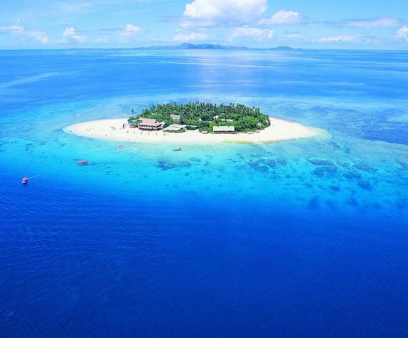 Aerial view of a small tropical island surrounded by vibrant turquoise and deep blue ocean. The island features lush greenery and sandy shores under a bright blue sky.