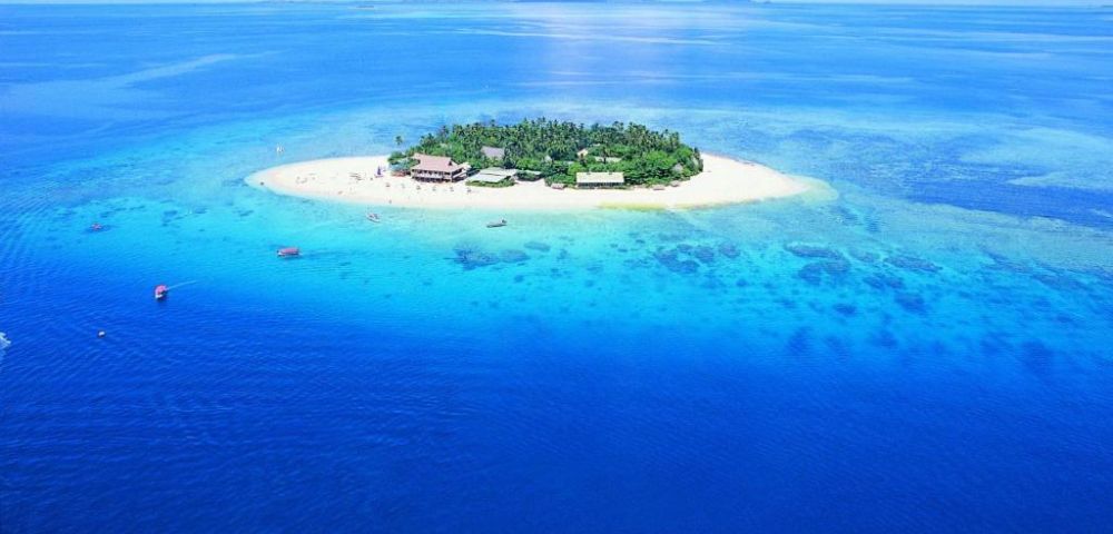 Aerial view of a small tropical island surrounded by vibrant turquoise and deep blue ocean. The island features lush greenery and sandy shores under a bright blue sky.