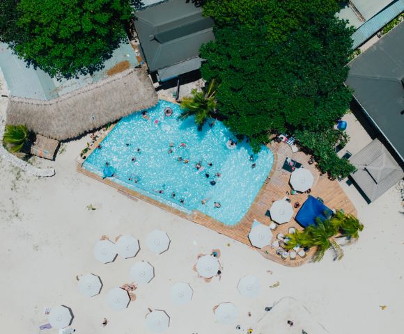 Aerial view of a beach resort with a pool. People swim and relax. Surrounding the pool are palm trees, sun umbrellas, and loungers on sandy ground.