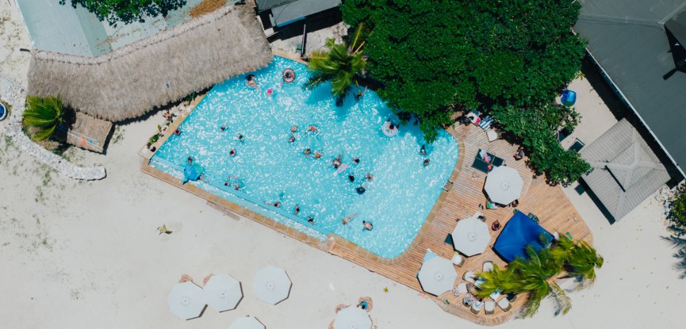 Aerial view of a beach resort with a pool. People swim and relax. Surrounding the pool are palm trees, sun umbrellas, and loungers on sandy ground.