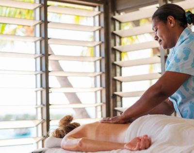 A serene image of a spa scene, showing a massage therapist in a blue uniform giving a back massage to a client on a bed, near sunlit louvered windows.