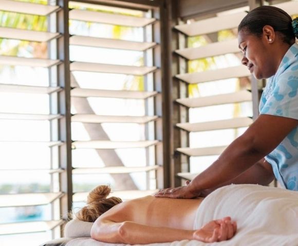 A serene image of a spa scene, showing a massage therapist in a blue uniform giving a back massage to a client on a bed, near sunlit louvered windows.