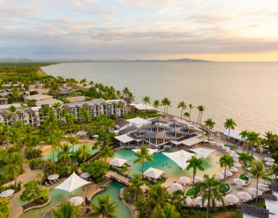 Aerial view of a tropical beachfront resort at sunset, with lush palm trees, turquoise pools, and modern buildings overlooking the calm ocean, evoking tranquility.