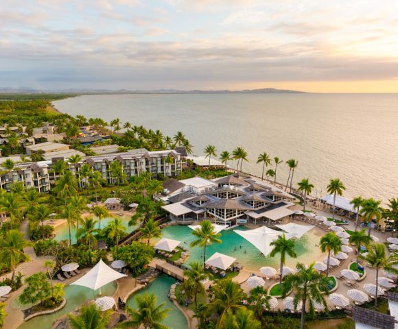 Aerial view of a tropical beachfront resort at sunset, with lush palm trees, turquoise pools, and modern buildings overlooking the calm ocean, evoking tranquility.
