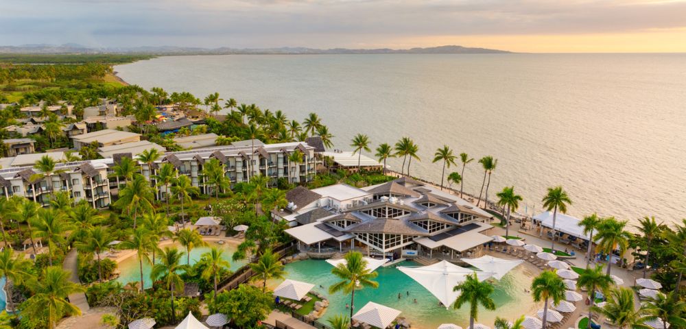 Aerial view of a tropical beachfront resort at sunset, with lush palm trees, turquoise pools, and modern buildings overlooking the calm ocean, evoking tranquility.