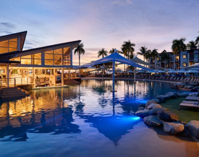 Modern resort pool area at sunset, with illuminated water, palm trees, and covered seating. Reflective, tranquil ambiance with warm lighting.