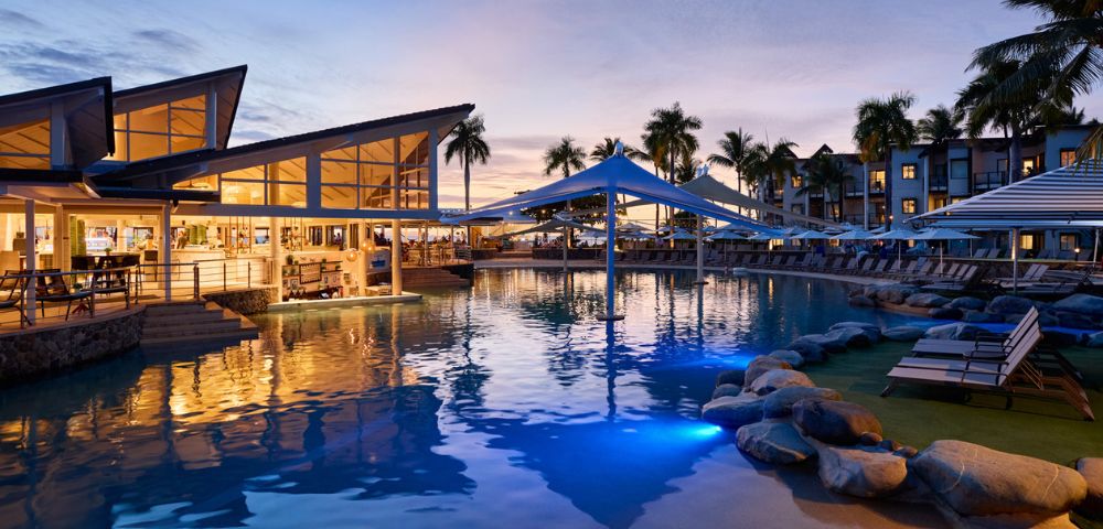 Modern resort pool area at sunset, with illuminated water, palm trees, and covered seating. Reflective, tranquil ambiance with warm lighting.