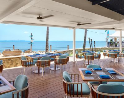 Outdoor beachside restaurant with wooden deck and blue cushioned chairs, overlooking calm ocean and clear blue sky. Relaxing and inviting atmosphere.