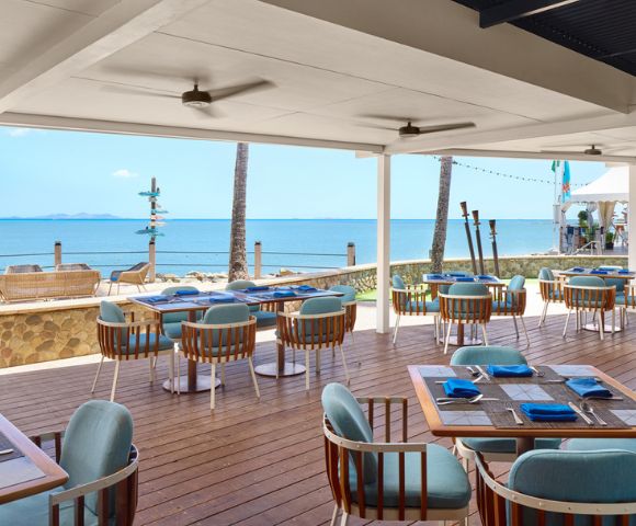 Outdoor beachside restaurant with wooden deck and blue cushioned chairs, overlooking calm ocean and clear blue sky. Relaxing and inviting atmosphere.