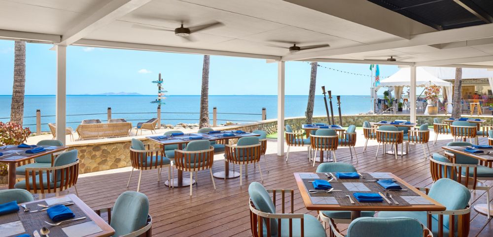 Outdoor beachside restaurant with wooden deck and blue cushioned chairs, overlooking calm ocean and clear blue sky. Relaxing and inviting atmosphere.