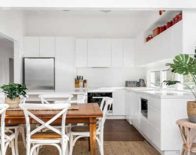 Bright, modern kitchen with white cabinets, stainless steel fridge, wooden dining table, white chairs, green plants, and red decor accents on shelves.